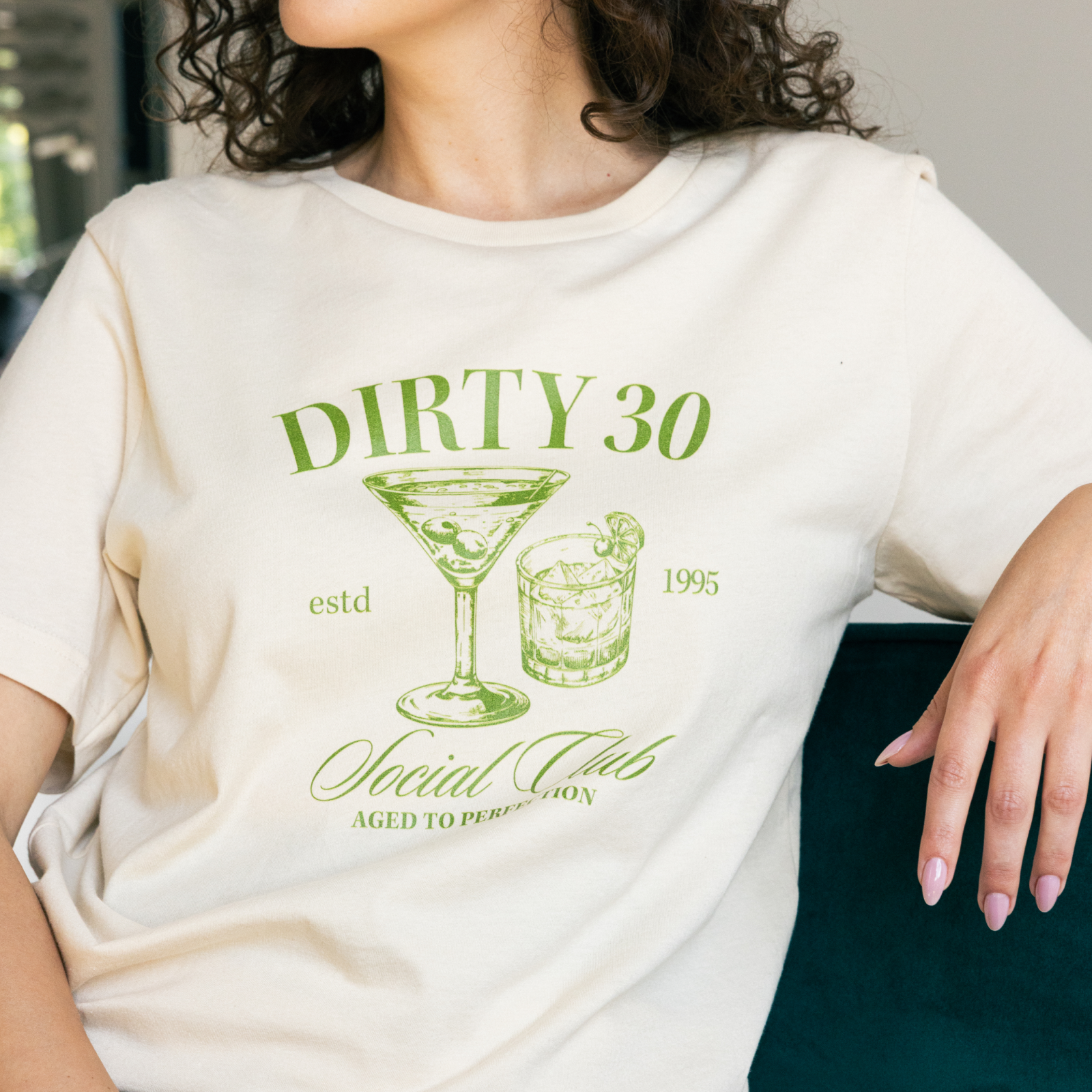 Woman wearing a beige t-shirt with green text and graphics reading "Dirty 30 Social Club," featuring cocktail images.