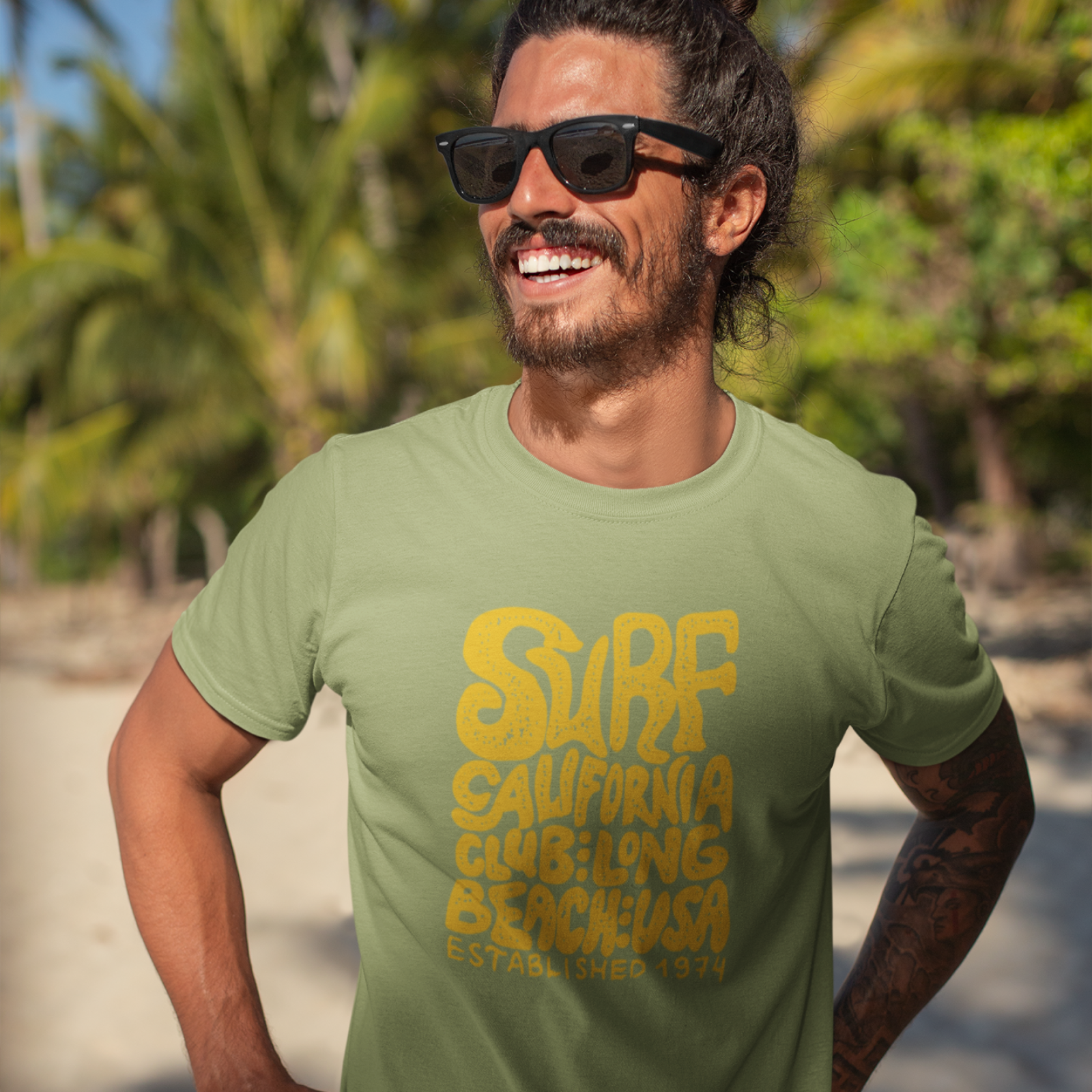 Smiling man with sunglasses and a beard, wearing a light green T-shirt that says "Surf California Club" in yellow.