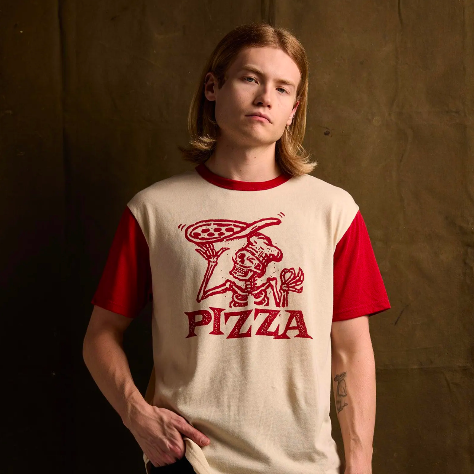 Young person with long hair wearing a beige and red t-shirt featuring a skeleton chef holding a pizza.