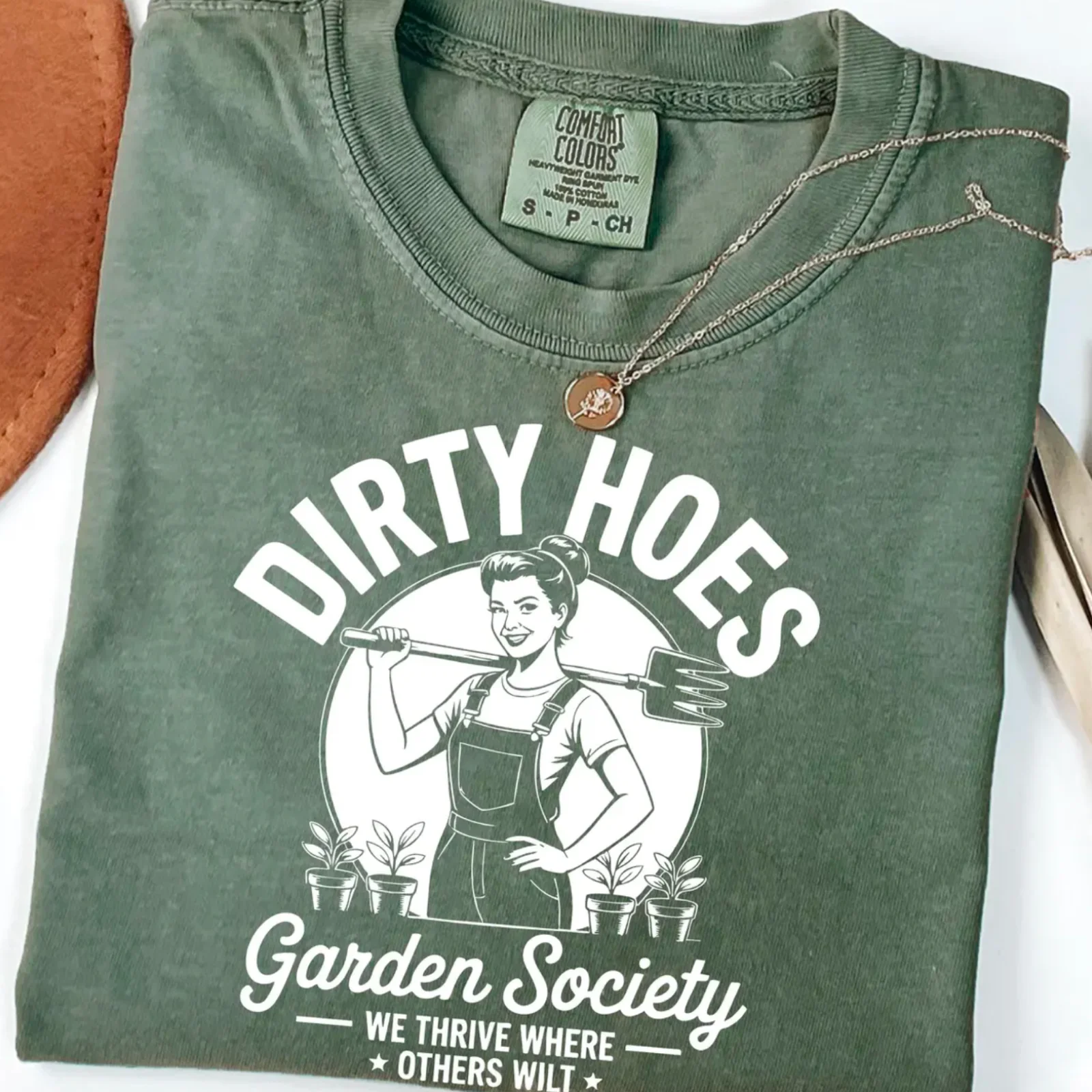 Folded green T-shirt with humorous text "Dirty Hoes Garden Society" and a vintage-style illustration of a woman with a pitchfork.