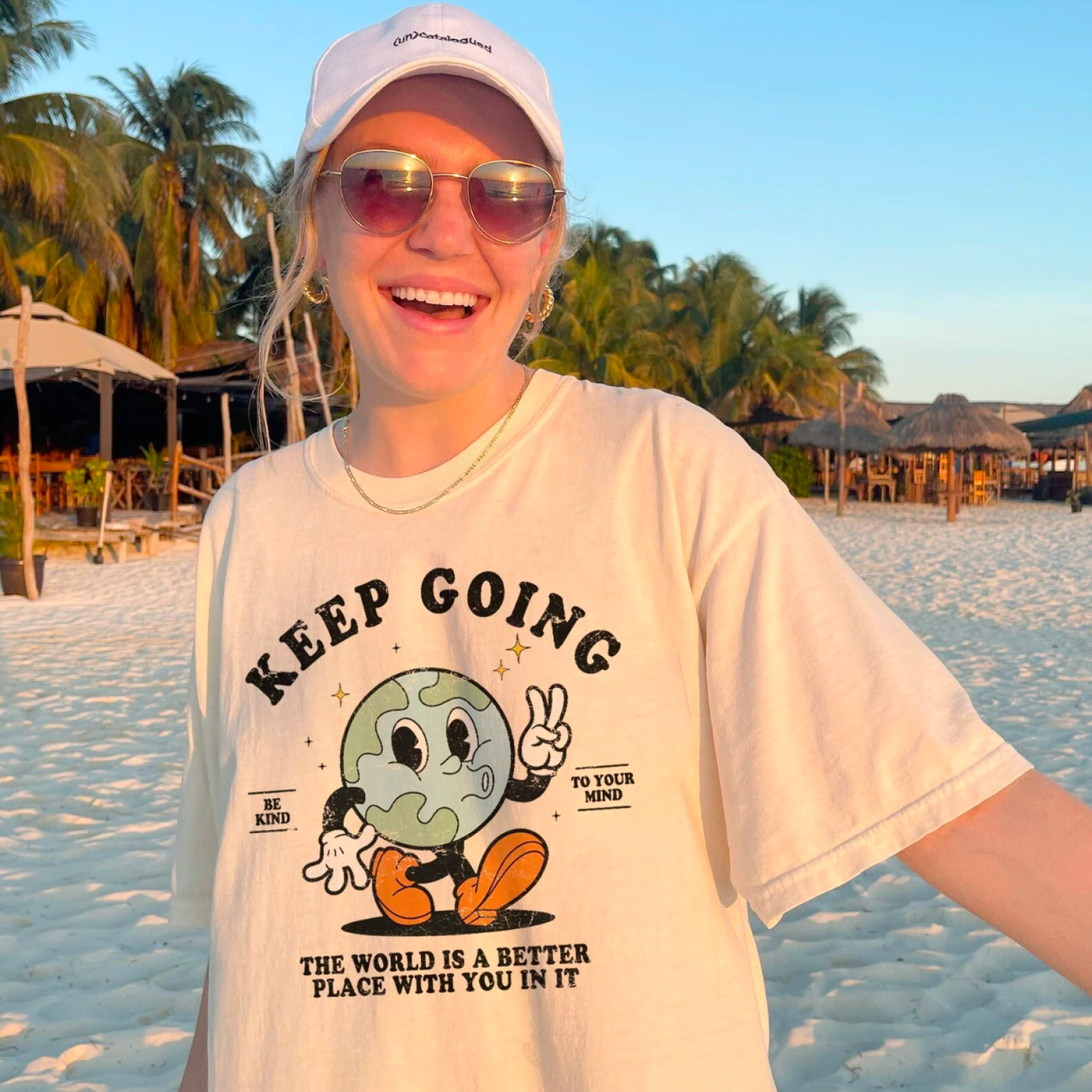 A smiling person in sunglasses and a white cap stands on a sunny beach.