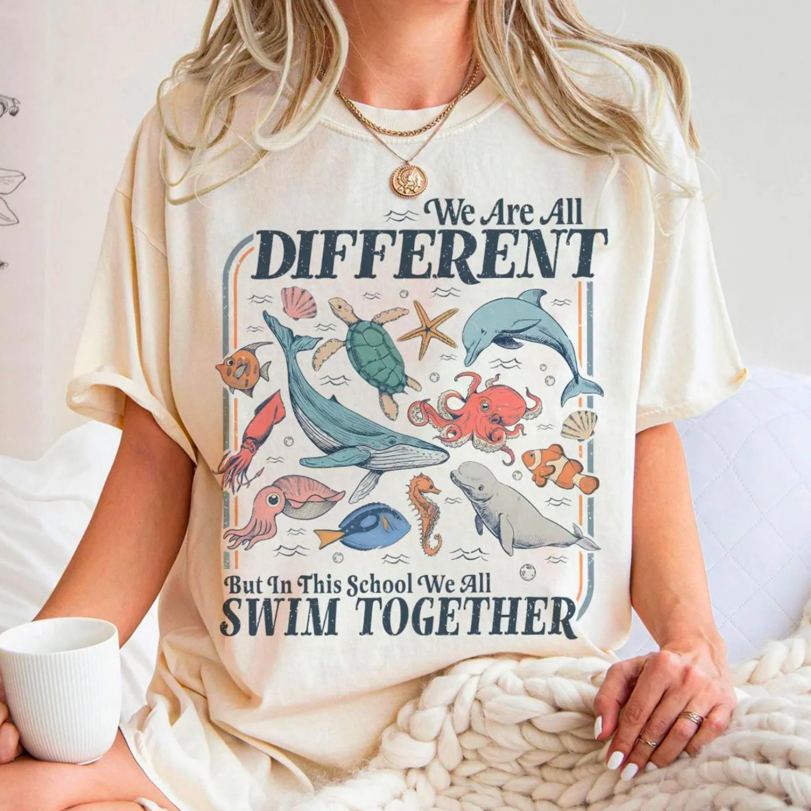 A woman wearing a light-colored t-shirt with marine animals and the words "We Are All Different But In This School We All Swim Together," while holding a mug.