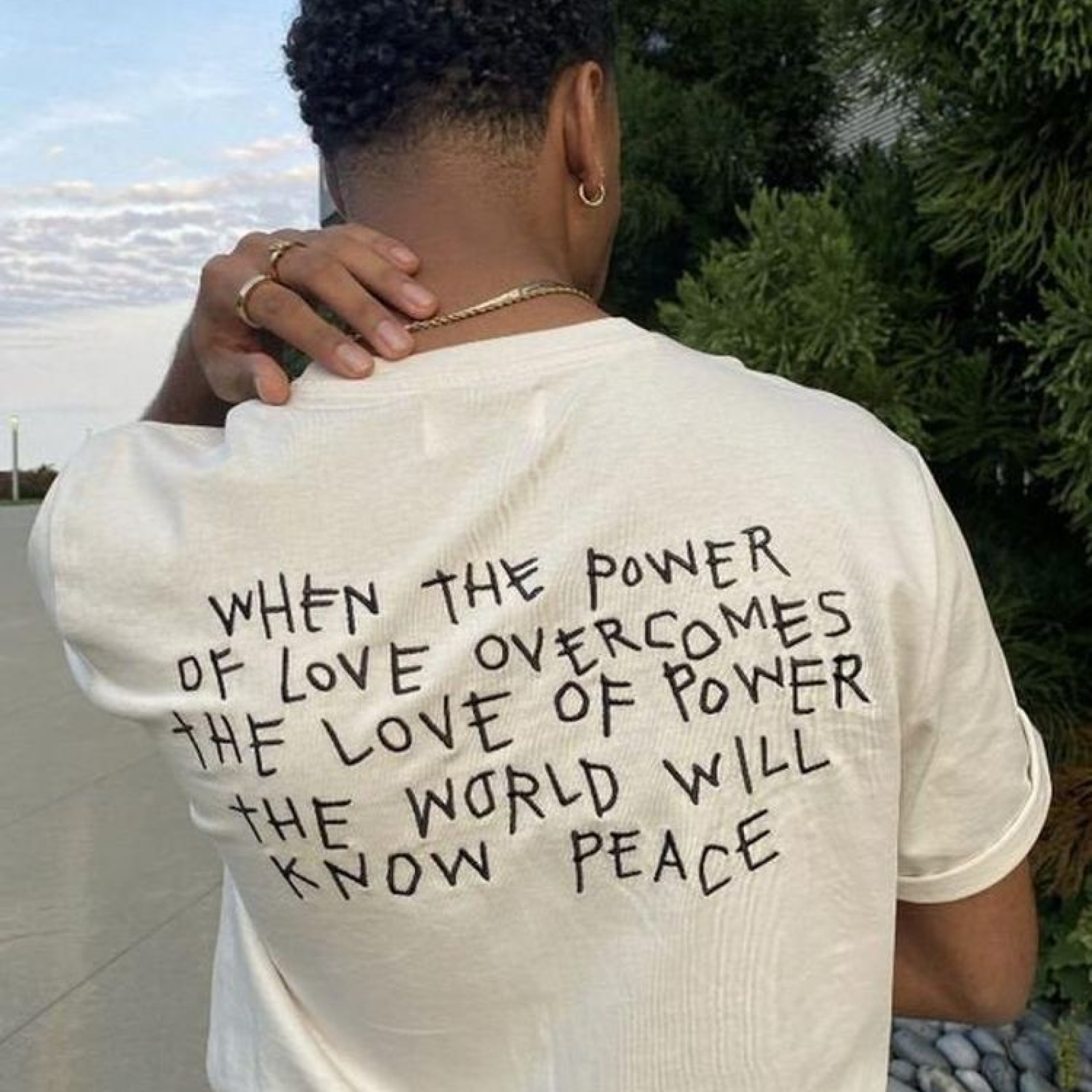A person with short curly hair wearing a white shirt with a handwritten quote about peace.
