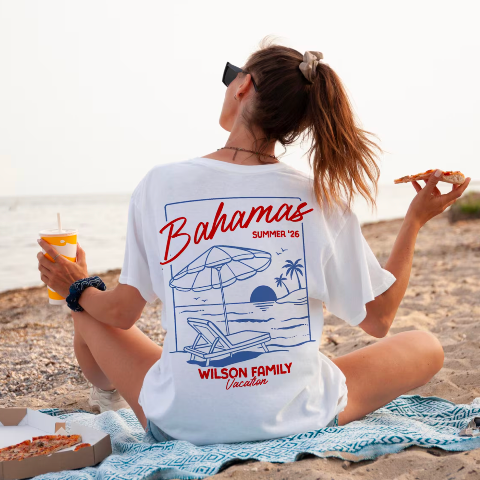 A woman sits on a beach, wearing a "Bahamas Summer '26" T-shirt, holding a drink and pizza slice.