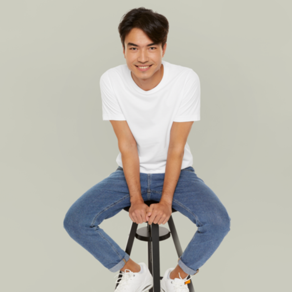 A man in a white t-shirt and jeans sits playfully on a stool against a gray background, smiling warmly at the camera, conveying a relaxed and cheerful mood.