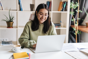 A woman in glasses and a green sweater smiles while working on a laptop at a bright desk.