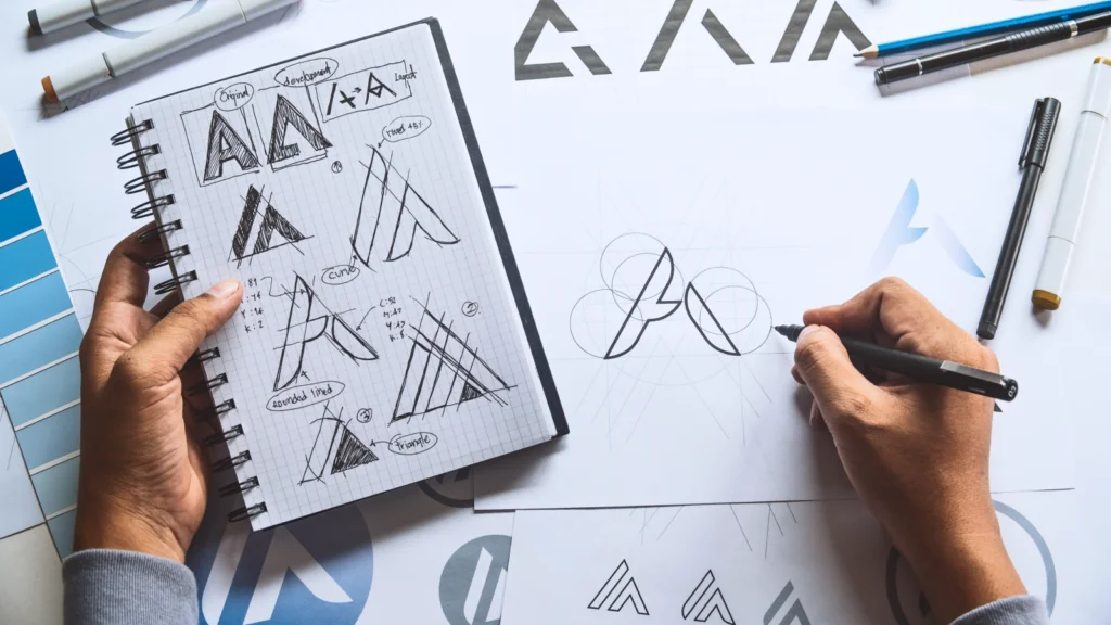 Hands drawing logo designs with geometric shapes on paper, surrounded by sketches and design tools.