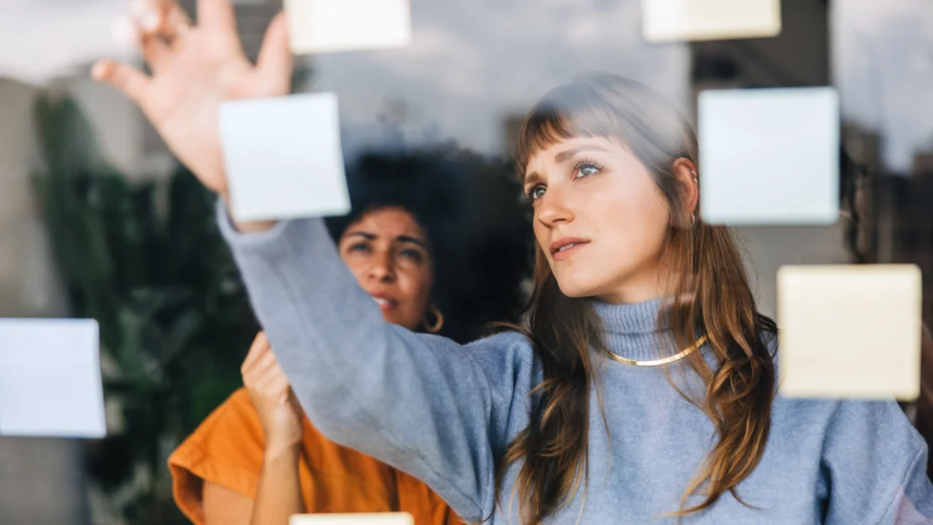 Two women brainstorming, looking at sticky notes on a glass wall.