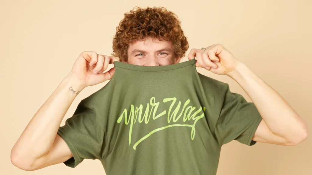 A person playfully lifts a green shirt with the words "your way" written in lime green script, partially hiding their face.