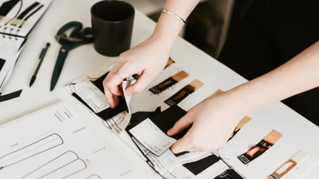 Hands work on a textile design project with fabric swatches, sketches, and scissors on a table, conveying creativity and focus.