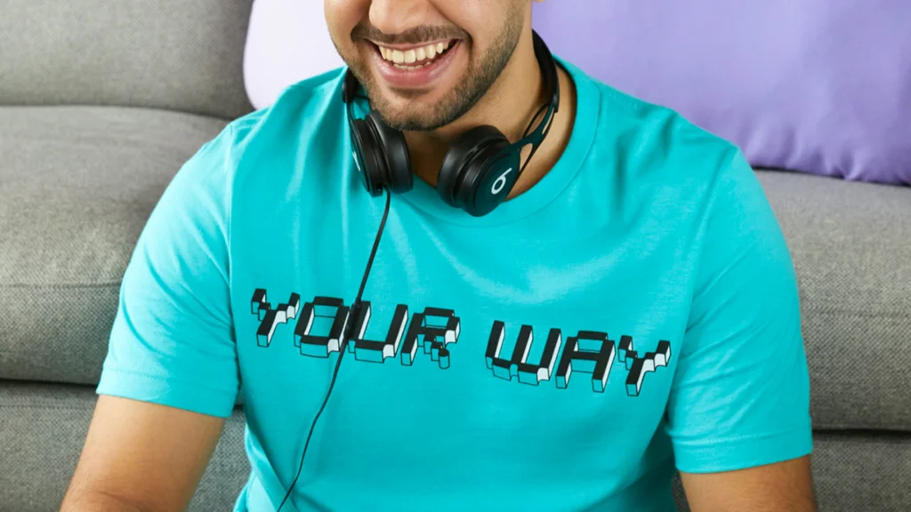 A smiling person wearing a turquoise "YOUR WAY" T-shirt sits on a gray sofa.