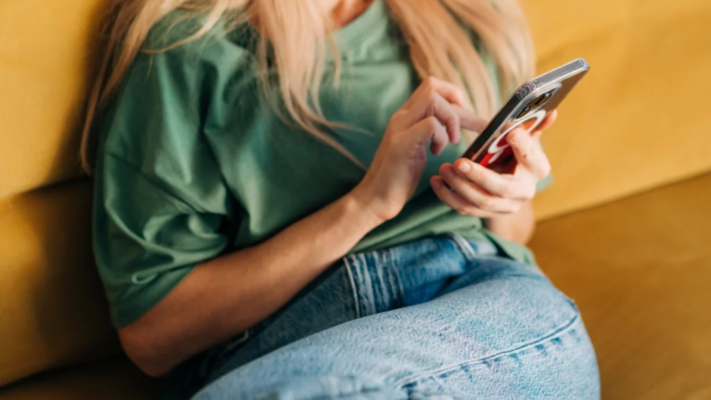 A woman in a green shirt and jeans sits on a yellow couch, using a smartphone, looking for the latest TikTok trends.