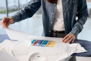 A person in a denim shirt applies a colorful "SUMMER" design onto a white T-shirt using heat transfer.