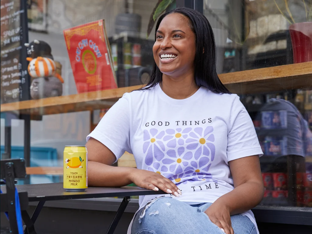 Picked your shirt Design and order it with Printify A woman smiles while seated outside a café, wearing a shirt that says "Good things take time." A can of mango milk rests on the table.