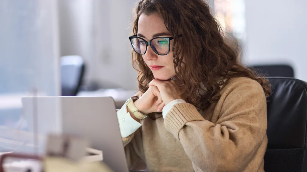 A woman with curly hair and glasses focuses intently on a laptop, learning how to design a logo. She's seated at a desk in a softly lit office.