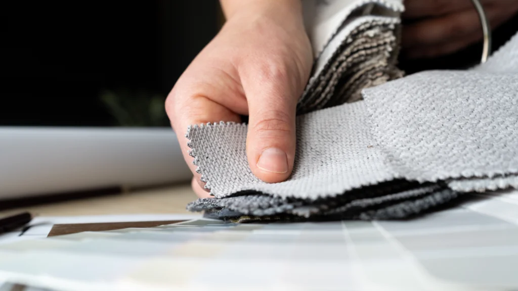 A hand flips through swatches of textured fabric in neutral tones on a table.