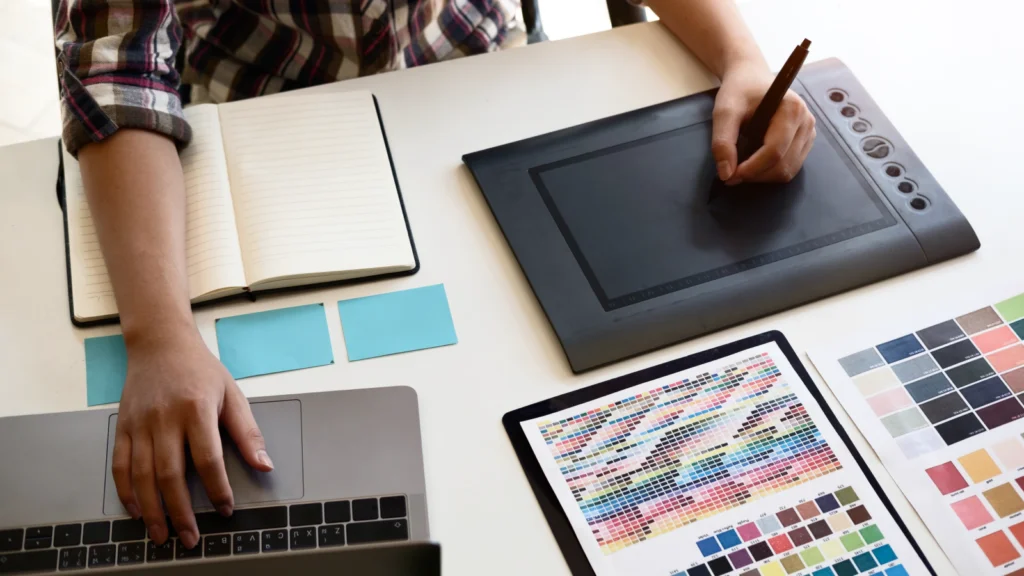 A person works at a desk with a drawing tablet, open notebook, and laptop.