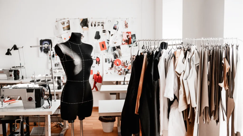 A fashion design studio with a black dress form, sewing machines, and a rack of neutral-toned garments.