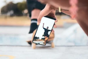 A person holding a phone captures a skateboarder performing a trick at a park. The skateboarder is mid-air.