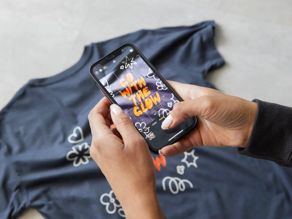 A person taking a picture of a dark-blue t-shirt with orange “Go with the glow” text, using a smartphone.