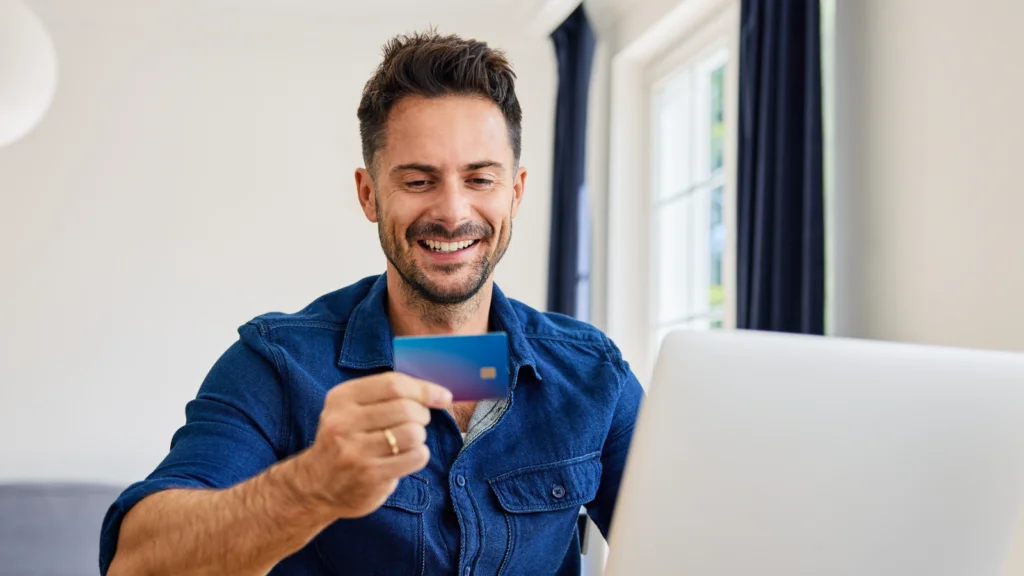A smiling man in a blue shirt holds a credit card while sitting at a laptop in a bright room, suggesting online shopping or banking.