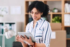 A woman in a striped shirt smiles while using a tablet in an office setting. Shelves with plants and books are in the background.
