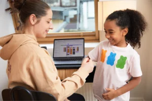 A smiling woman sits at a desk with a laptop showing a planner. She interacts happily with a child wearing a shirt with colorful bear shapes.