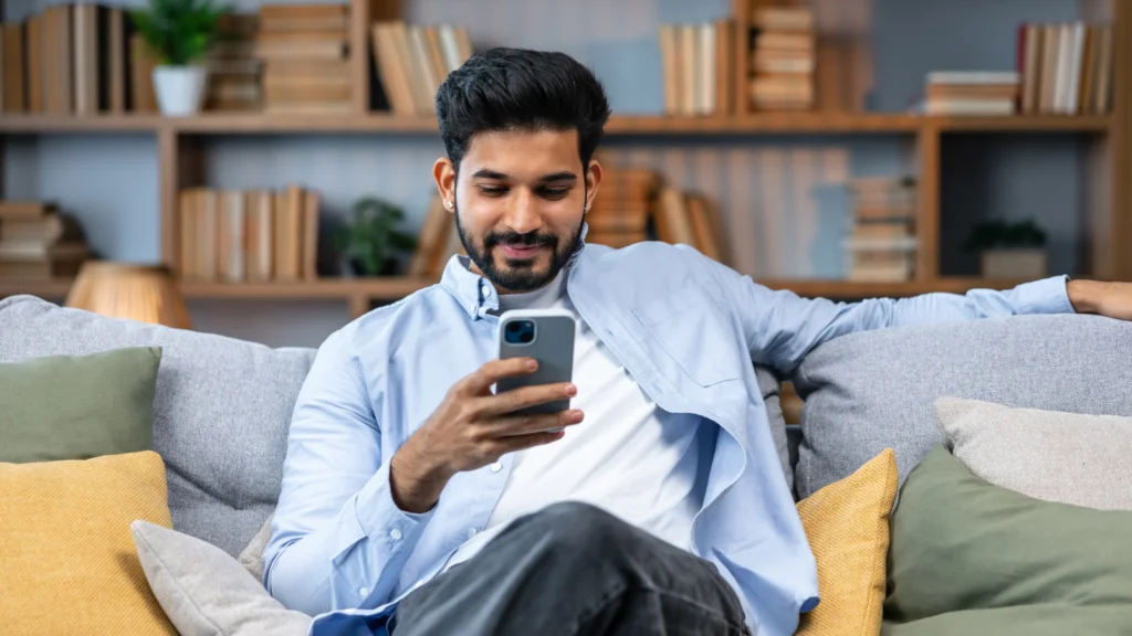 01 Optimize your profile for conversions A man relaxes on a couch, smiling while looking at his smartphone. He's wearing a light blue shirt, with cozy pillows around him.