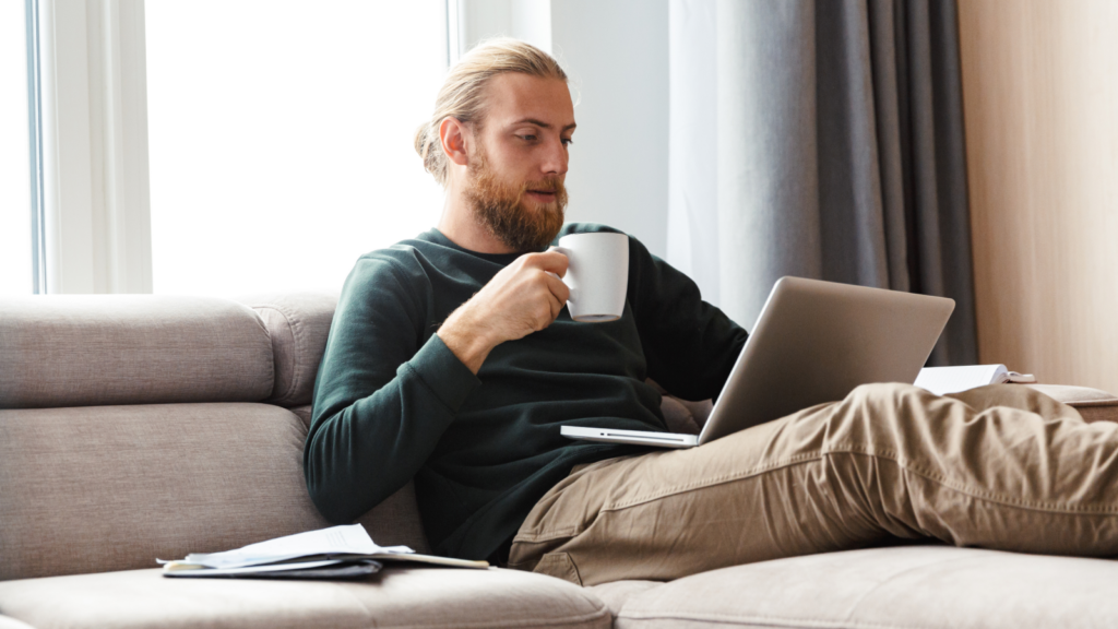 A bearded man is sitting on a couch with his legs up, drinking coffee and looking at a laptop.