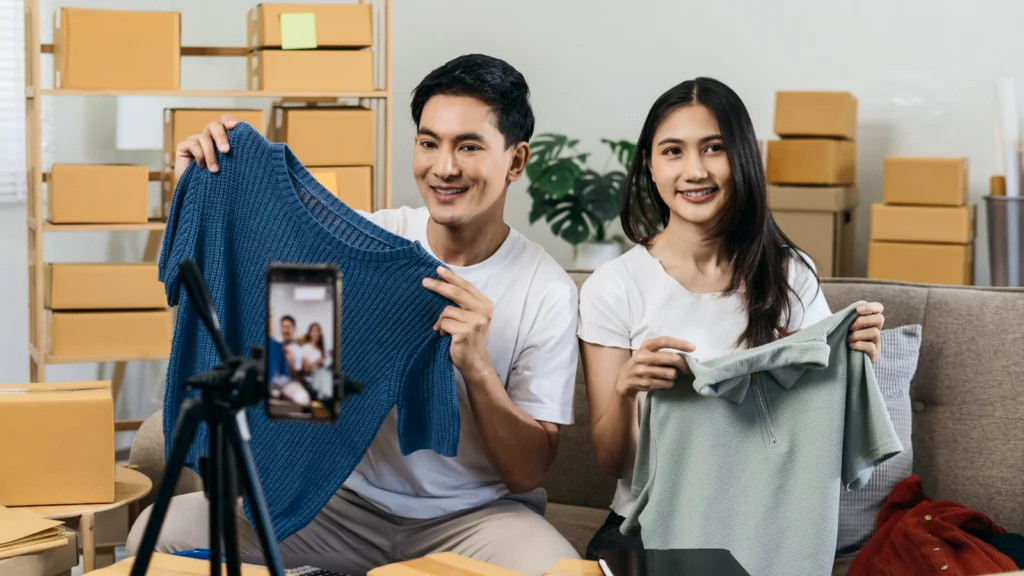02 Use TikTok LIVE to sell your products A smiling man and woman hold shirts while live-streaming on a smartphone in a room filled with cardboard boxes.