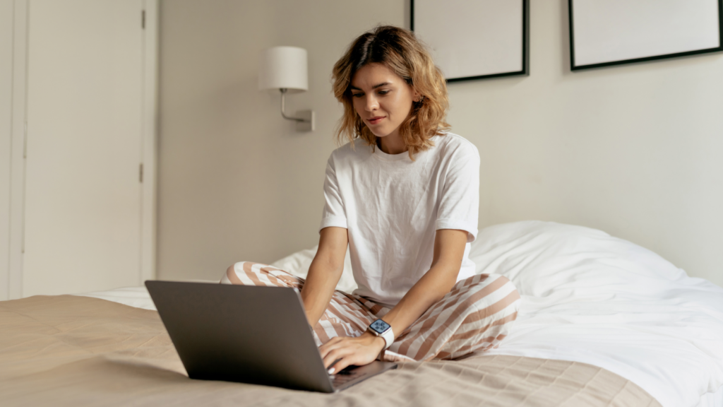 03 Create your Amazon account A woman is sitting on top of a bed, typing on a laptop in front of her, searching for how to sell Print on Demand on Amazon.