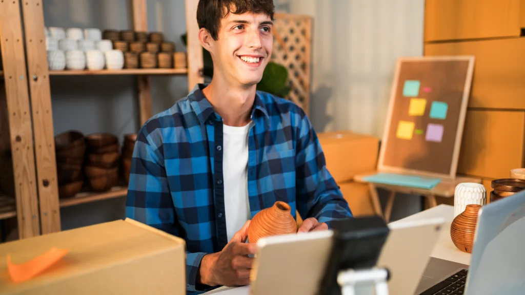 03 Promote your products with affiliates A smiling man holding a ceramic vase, sitting at a cluttered work desk with a laptop and packages. Pottery and a board with notes behind him.