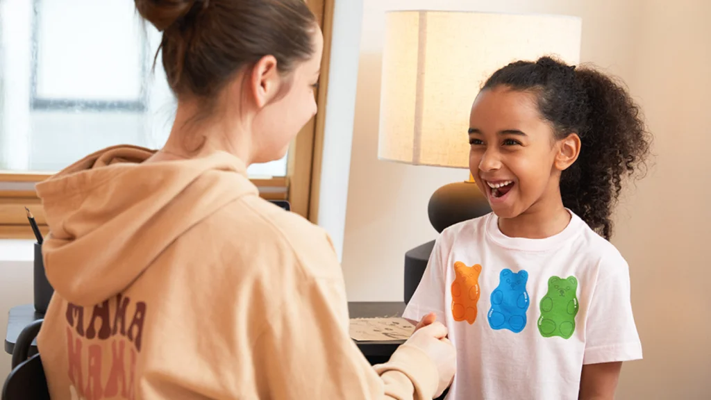 A woman in a tan hoodie smiles at a laughing girl wearing a white shirt with colorful bear designs.