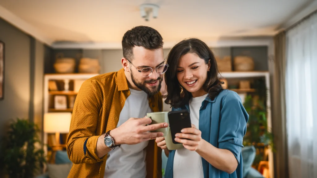 06 Common TikTok marketing mistakes A smiling couple in a cozy living room looks at a smartphone together. The man holds a mug. Shelves in the background.
