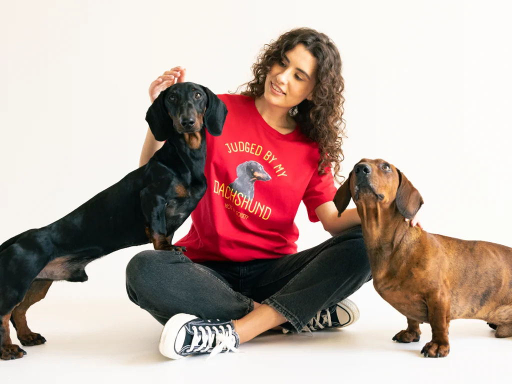A woman in a red shirt with a dachshund graphic sits cross-legged, smiling at two dachshunds.