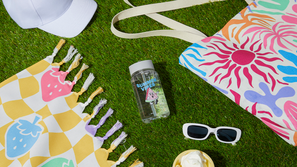 1 7 Beach essentials for hot summer days Summer-themed items on grass: a patterned towel with fruit, a vibrant sun tote, a water bottle, white cap, sunglasses, and ice cream cone.