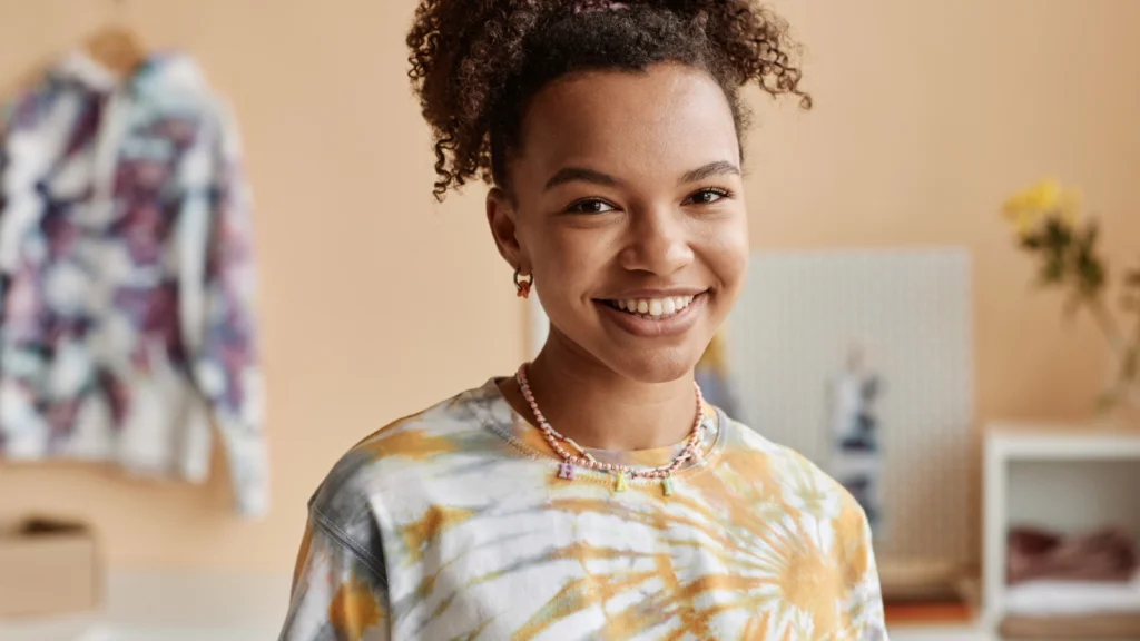 Smiling person in a tie-dye shirt and necklace stands in a cozy room.