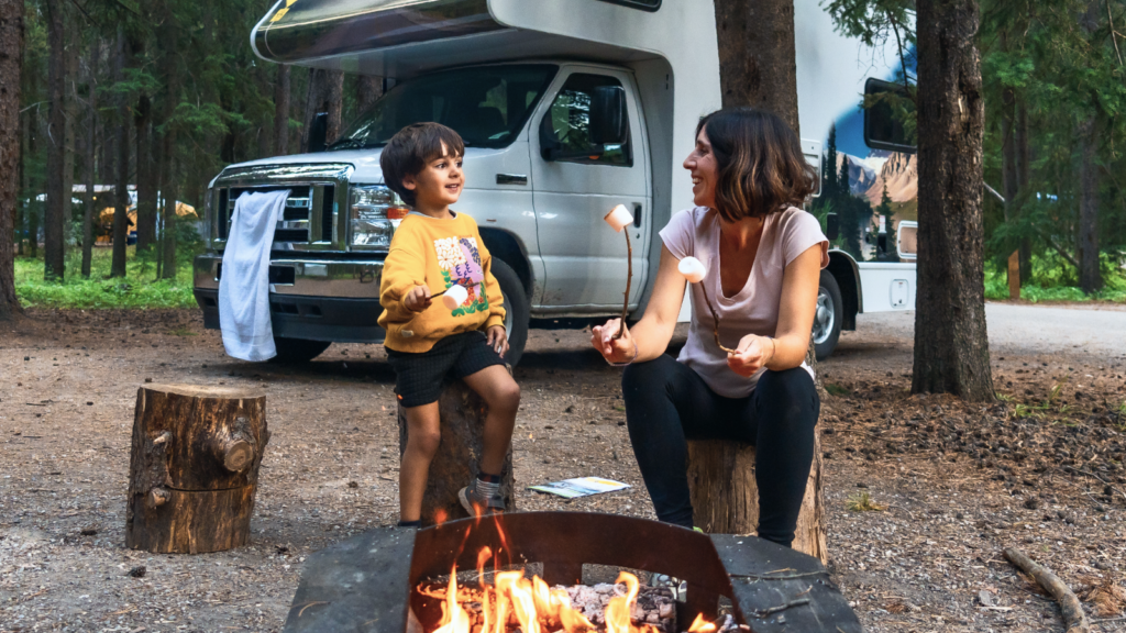 5 9 Summer outdoor gear for campers and travelers A woman and child toast marshmallows over a campfire in a forest, near an RV.