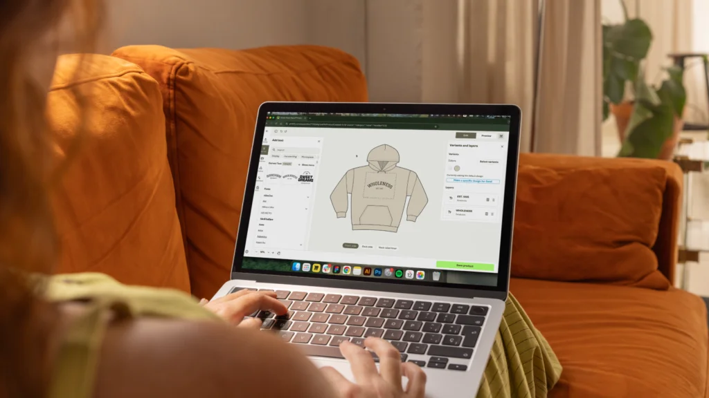Add your design and customize the products A woman sitting on an orange couch, using a laptop to design a beige hoodie with Printify’s Product Creator.