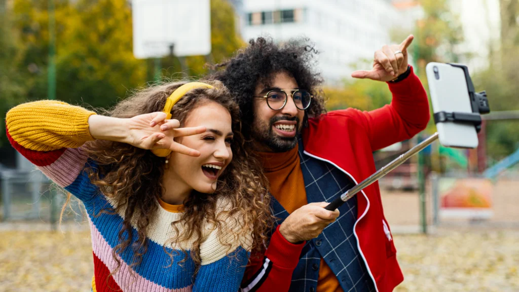 Collaborate with other creators A joyful young couple in colorful sweaters takes a selfie with a phone on a selfie stick, both smiling widely.