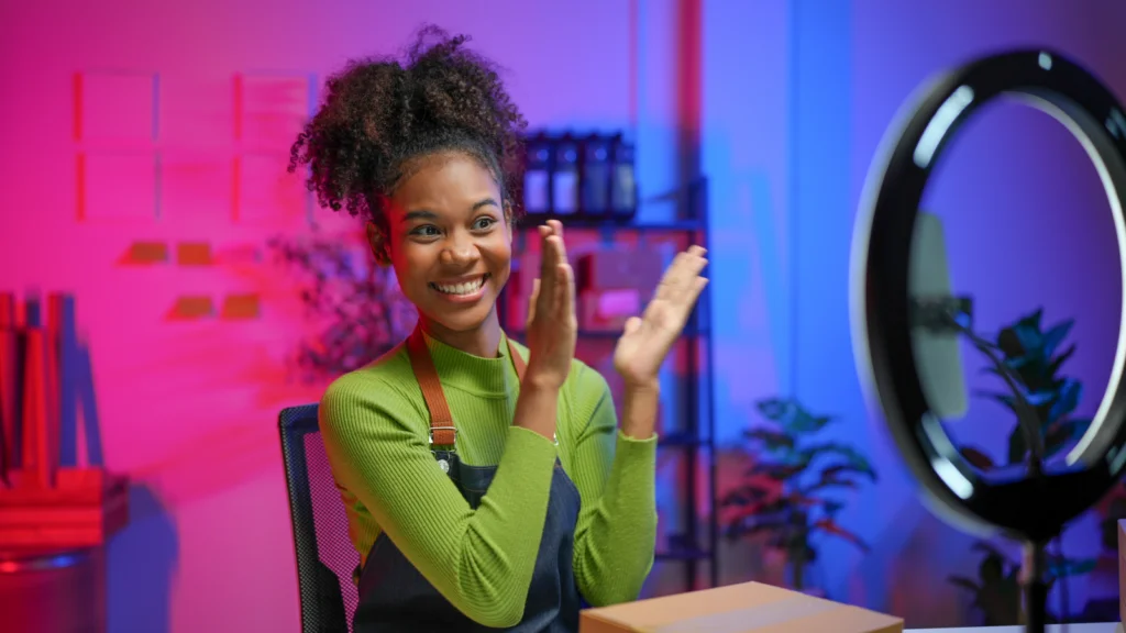 A smiling woman in a green sweater and apron claps happily while live-streaming in a colorful room with pink and blue lighting and plants.
