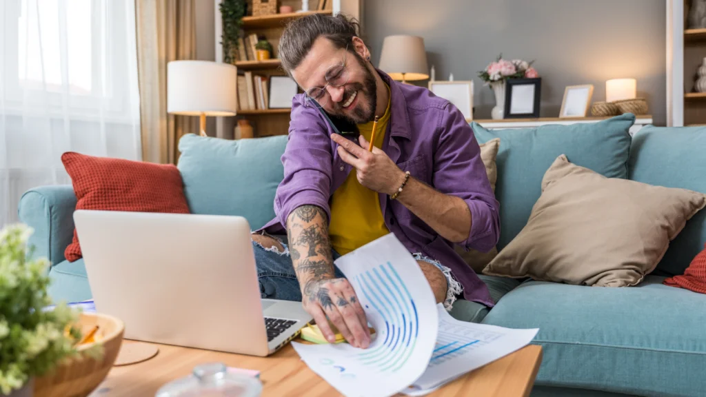 How TikTok analytics works A cheerful man with tattoos talks on a phone, sitting on a cozy sofa. He's reviewing paperwork by a laptop.