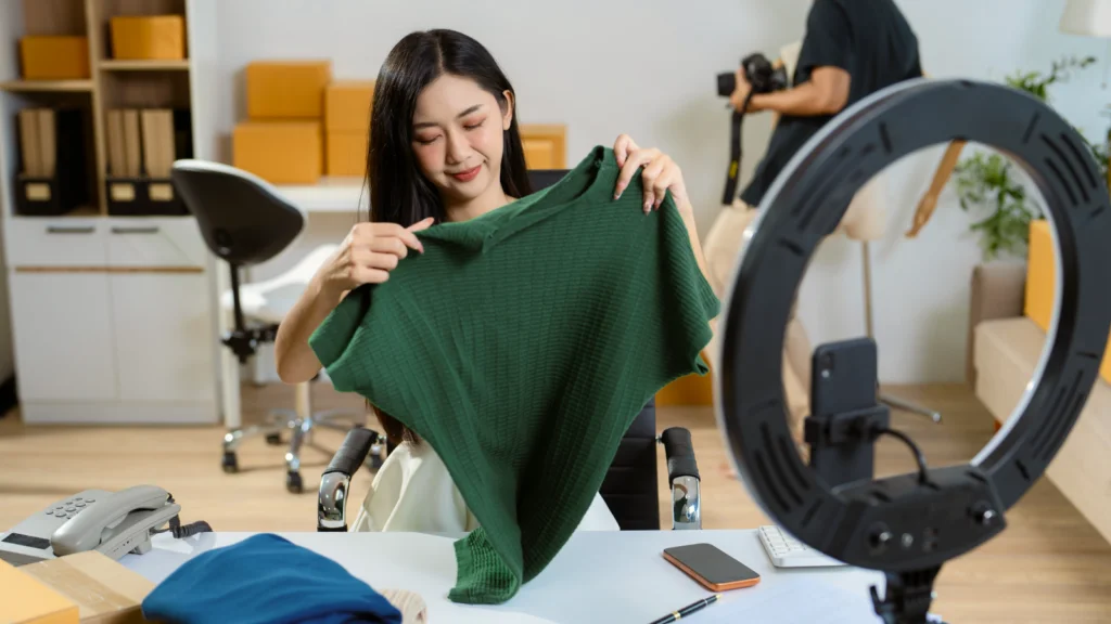How to improve TikTok engagement A woman smiles while holding a green shirt in front of a camera setup with a ring light, as if reviewing or showcasing the item.