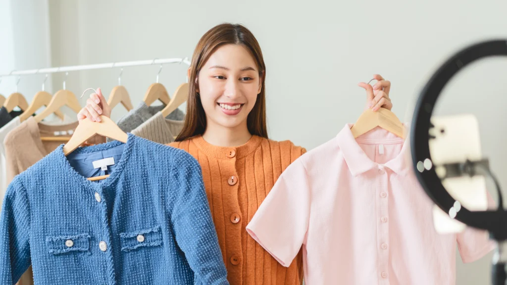 How to pitch TikTok creators effectively A woman is holding two pieces of clothing (blouse and polo shirt) in front of a smartphone on a stand with a ring light.