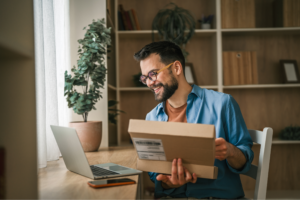 A man with glasses and a beard sits at a desk, working on a laptop beside an open box.