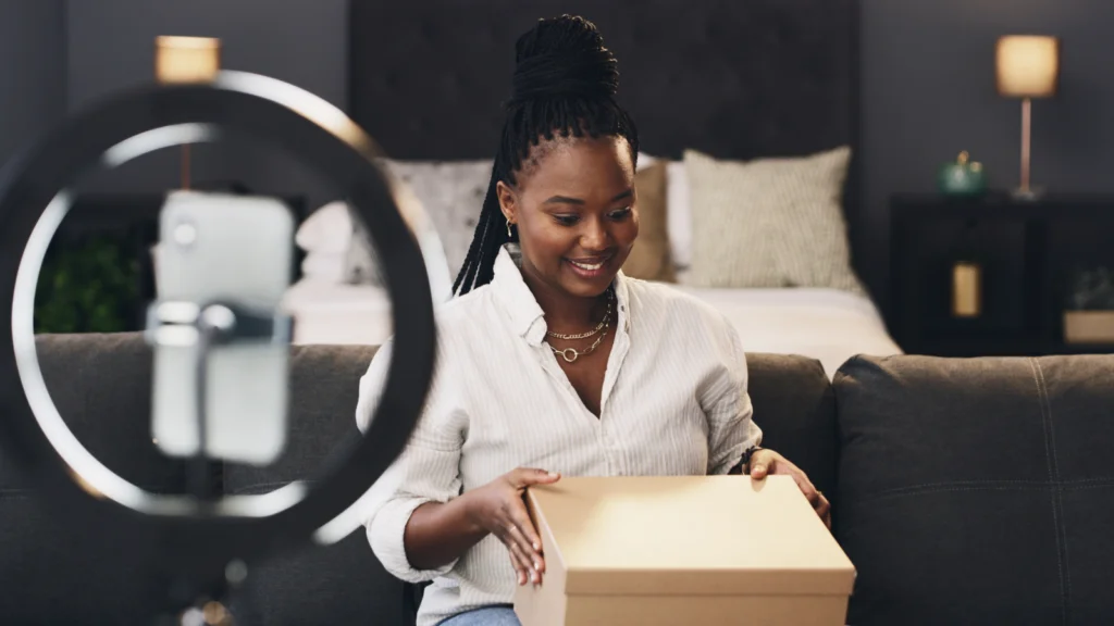 Maintain a strong Product Rating A woman is unpacking a delivery box during a livestream, using a smartphone on a stand with a ring light.