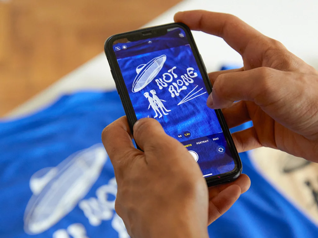 Monetize your TikTok followers with Printify A person holds a smartphone capturing a photo of a blue shirt with a UFO and aliens, featuring the text "Not Alone”.