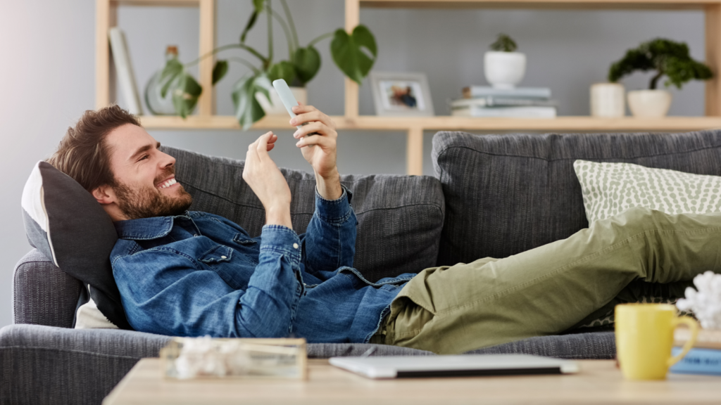 Post for volume first then optimize A man in a denim shirt and khaki pants lounges on a gray sofa, smiling at his smartphone.