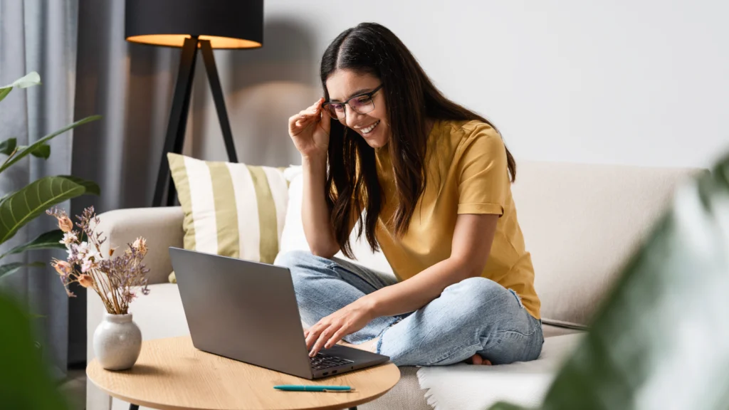 Printifys Pop Up Store can be your key to success A woman wearing glasses sits cross-legged on a couch, smiling as she uses a laptop to learn how to create a Printify Pop-Up Store.