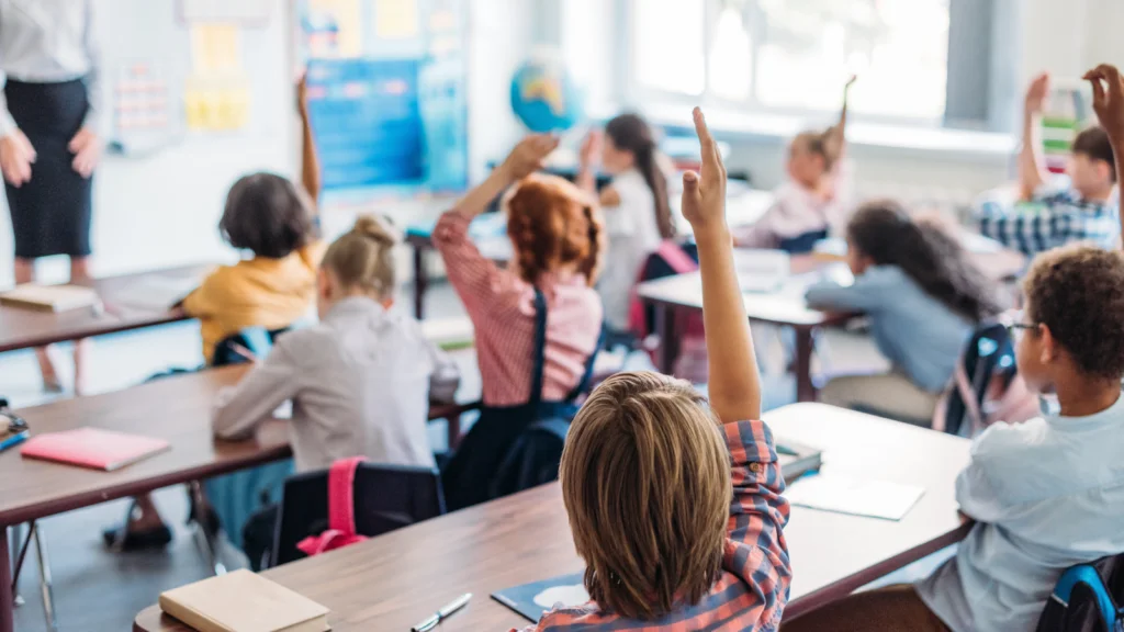 Q3 The back to school rush A classroom with young students sitting at desks, raising their hands eagerly.